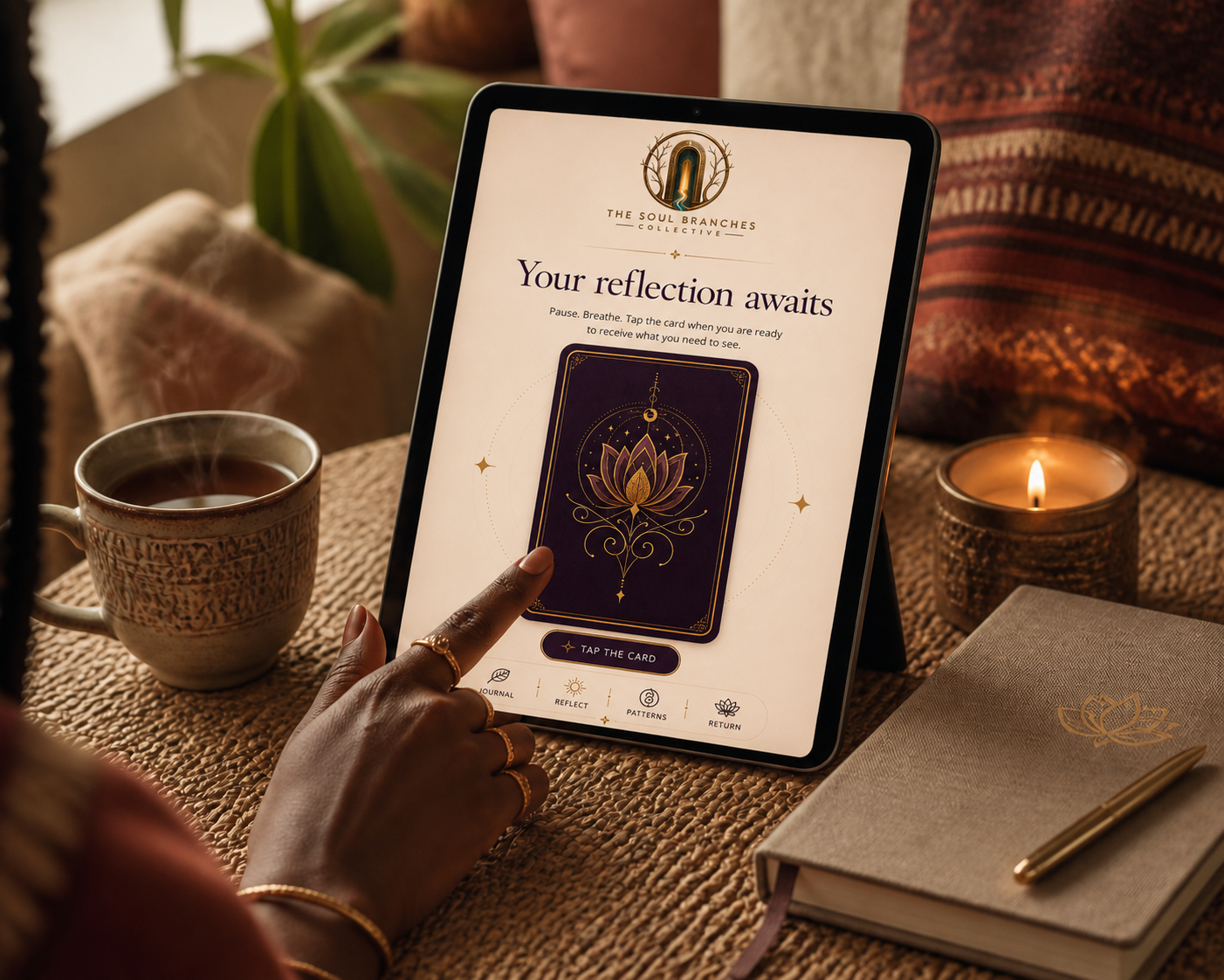 A tablet on a woven table showing a cream-and-plum Soul Branches reflection card, touched by a Black woman's hand, beside a tea cup, lit candle, and journal.